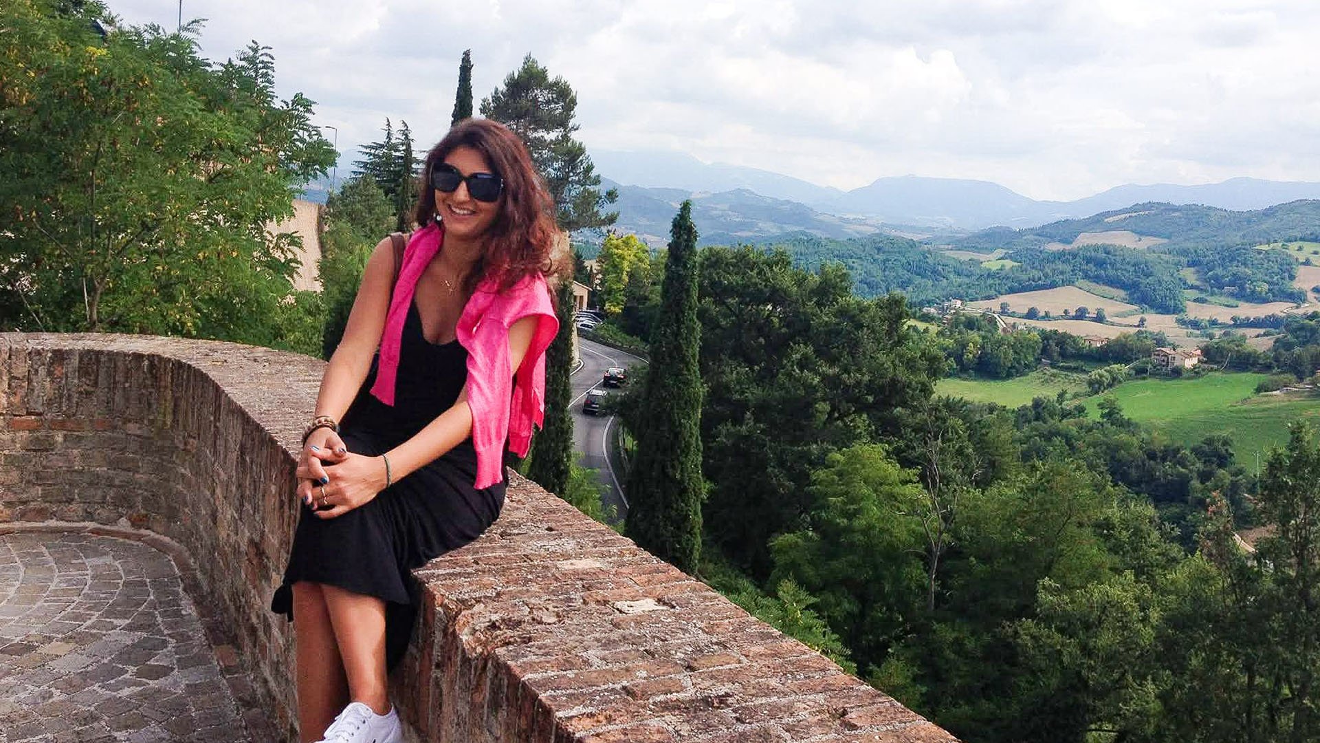 A woman sits on the edge of a brick wall against a backdrop of rolling hills in Urbino, Italy