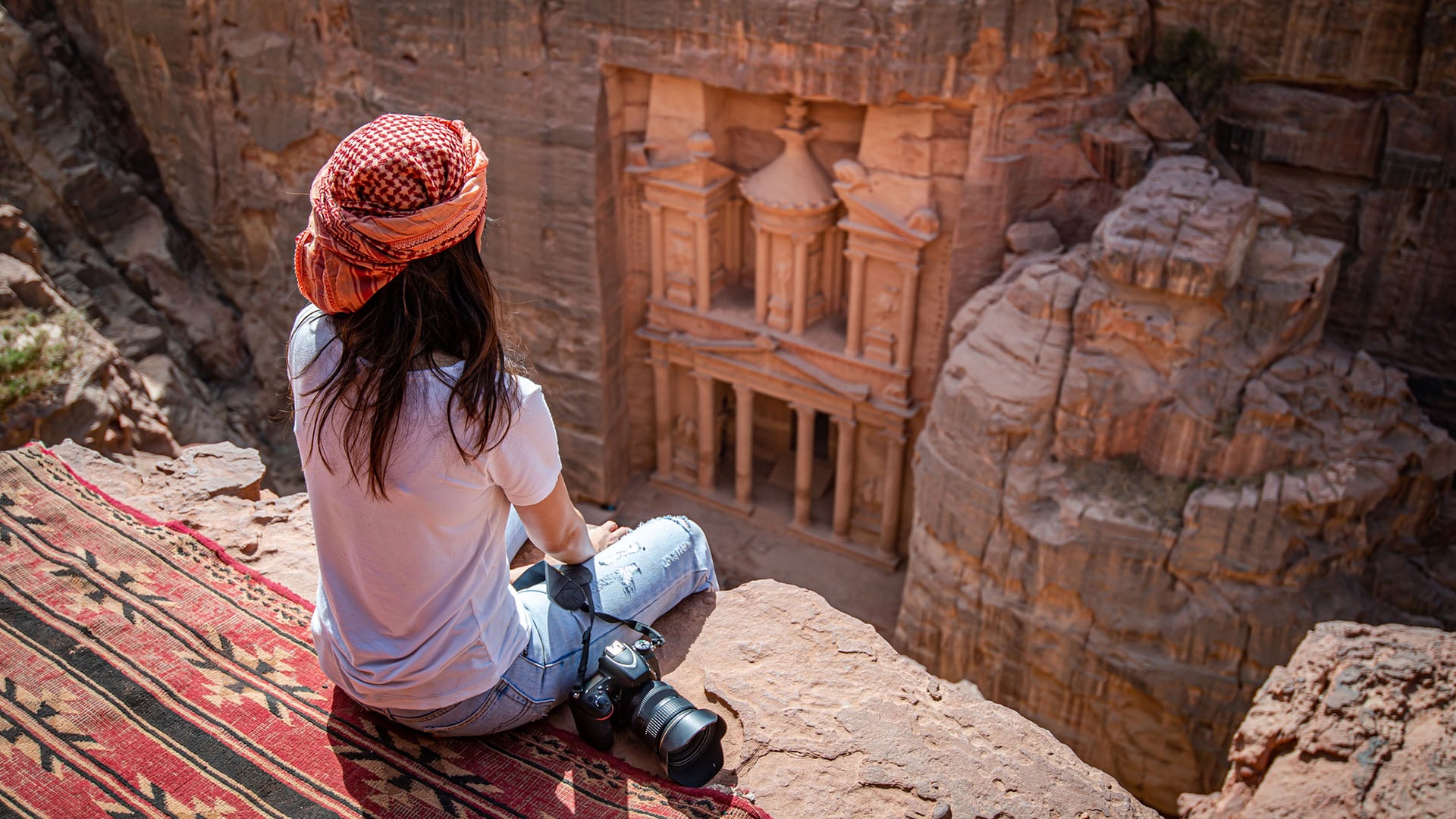 A woman with a camera sitting on a rocky overhang looking down on Petra's Treasury in Jordan
