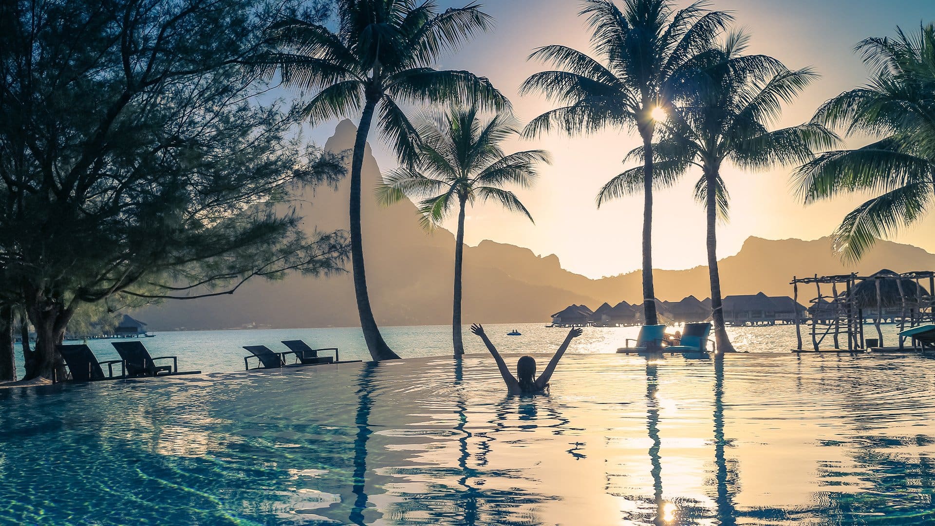 person in an infinity pool looking at ocean with overwater bungalows and a mountain silhouette