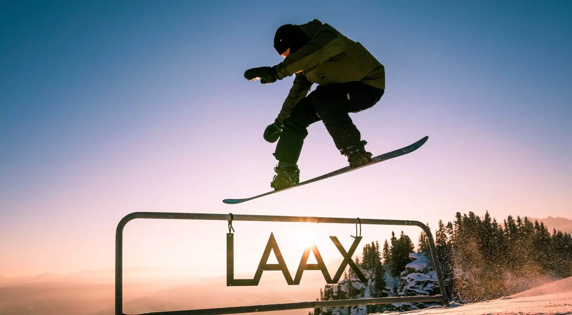 A snowboarder jumping over a sign saying "Laax" at sunset, in Laax, Switzerland.