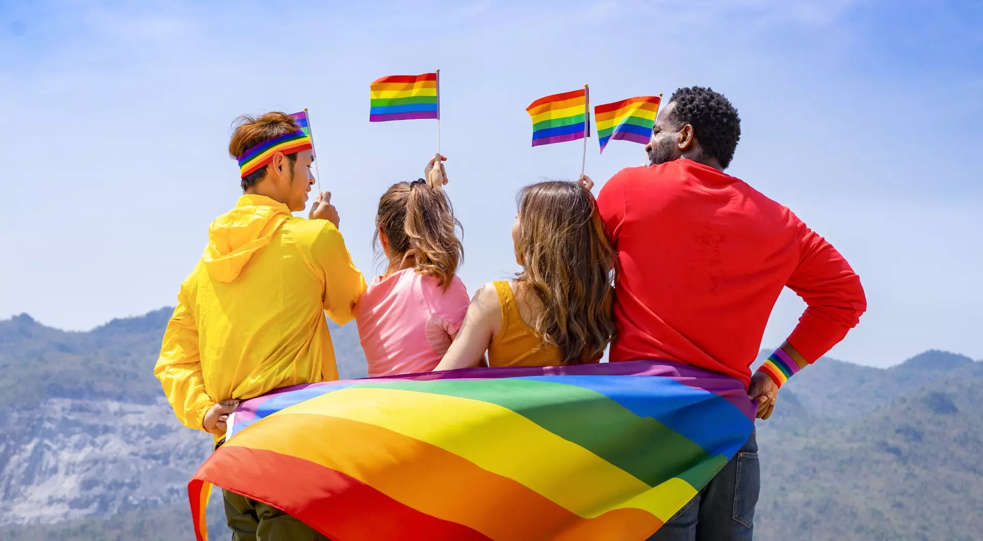 Group of diverse lgbtq friends wearing colorful clothes, standing side by side, smiling and holding lgbt flags waving in the air