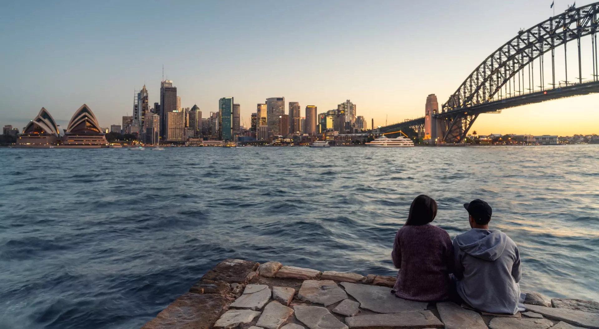 Sydney Harbour, Sydney, Australia couple sitting on edge of harbour looking at bridge and skyline