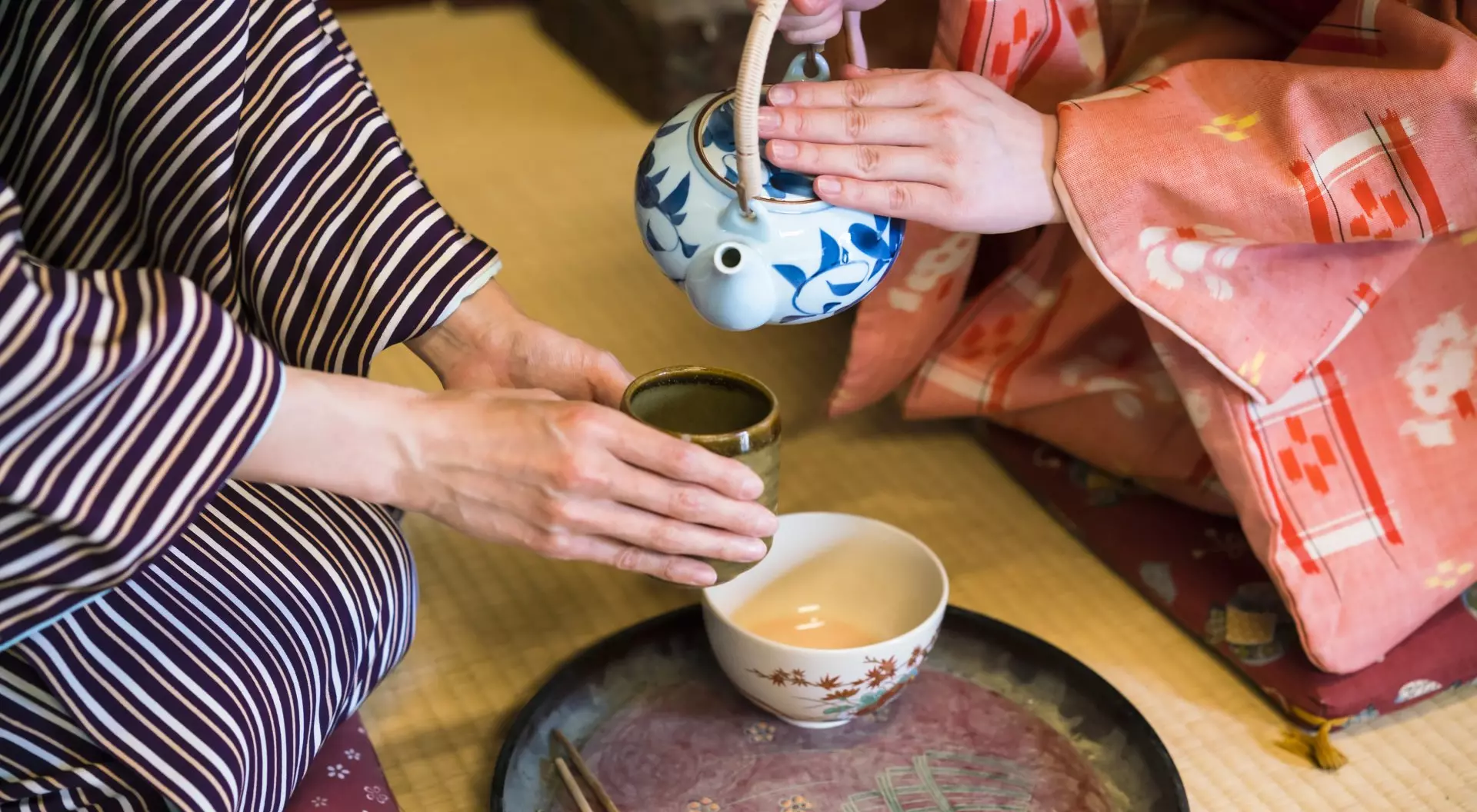 Japan detail of two women in traditional kimono, kneeling on tatami preparing, pouring a cup of tea which is inthe hands of one woman. They are in traditional Japanese old house on tatami. This is in Toei studios in Kyoto with old buildings from Samurai times.