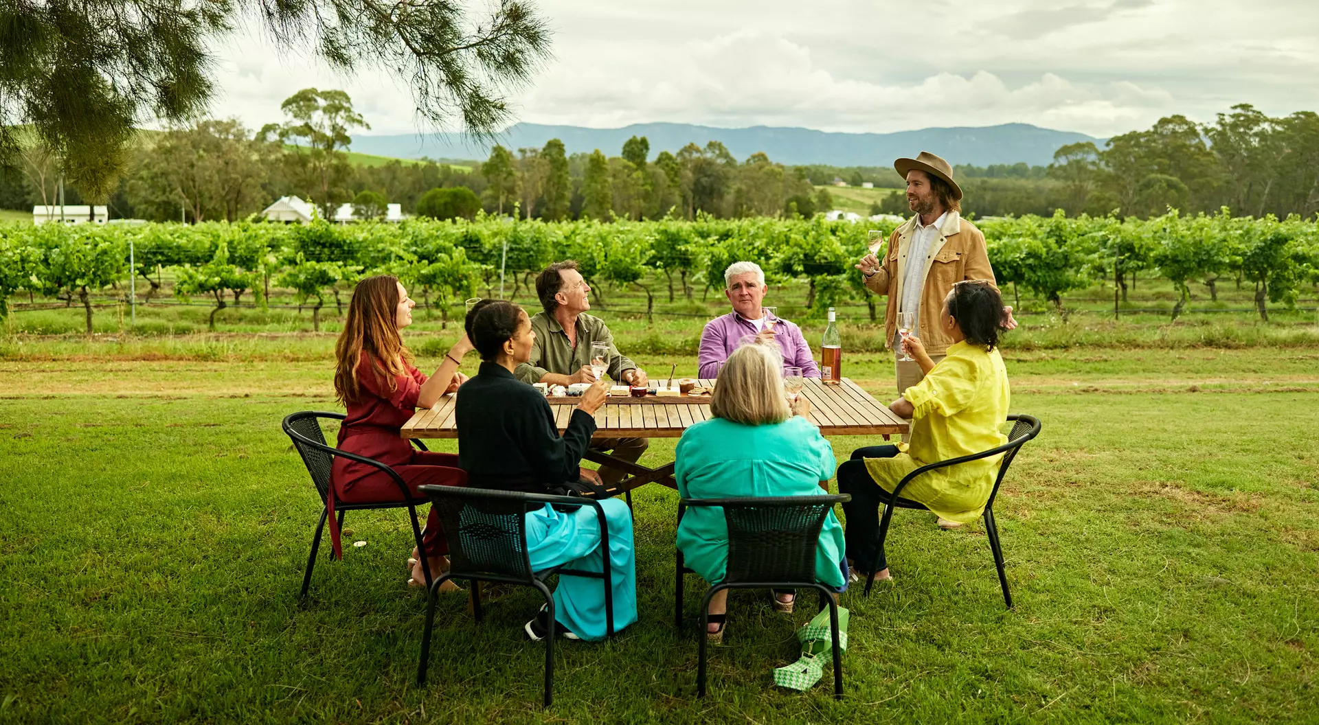 A group of six travellers sharing a toast with wine glasses at a table outdoors, with a vibrant green vineyard in the background and a green tree hanging overhead.