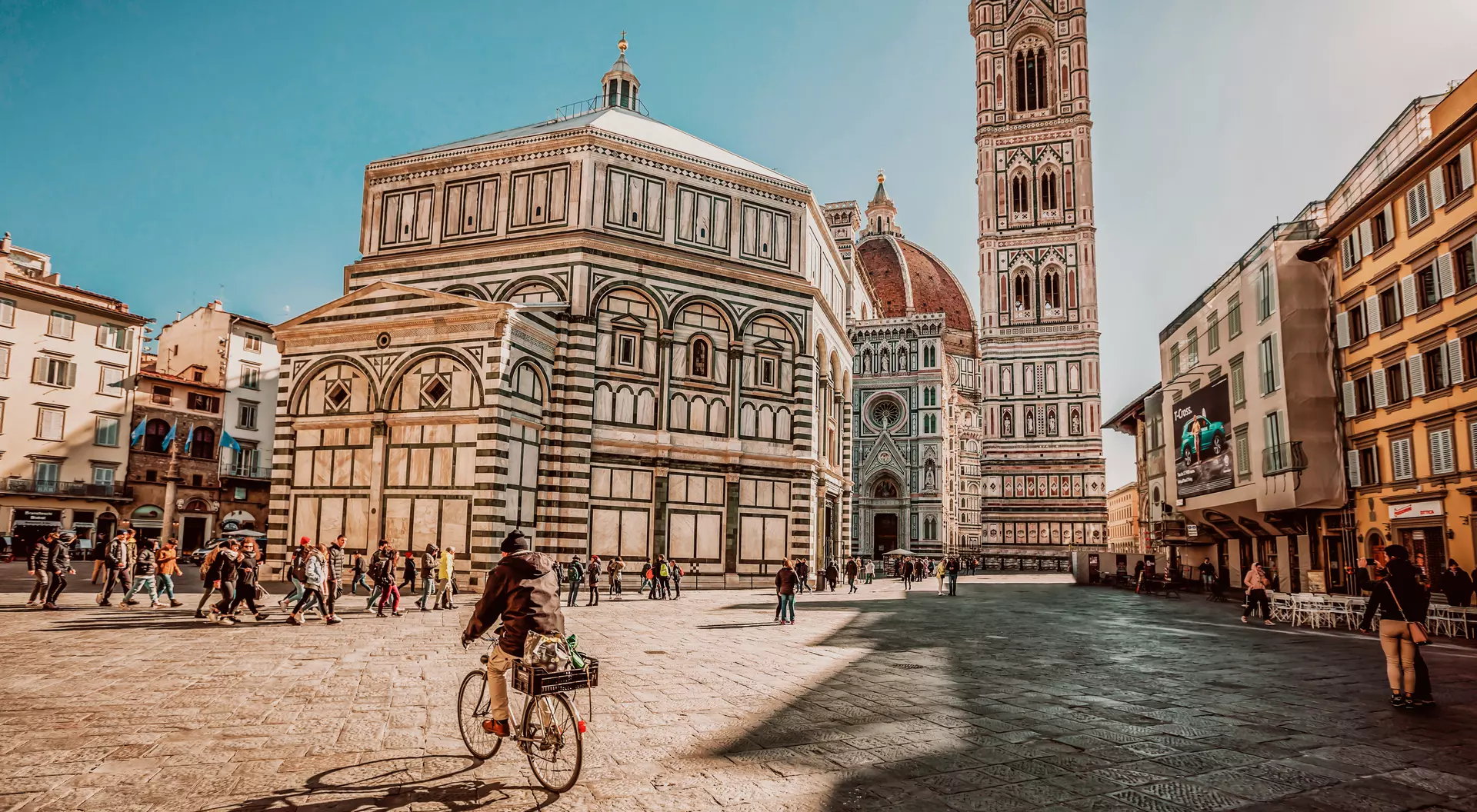 a man rides a bike into the charming old square of Piaza del Duomo in Florence, where the massive cathedral looms in the background with an ever-present crowd.