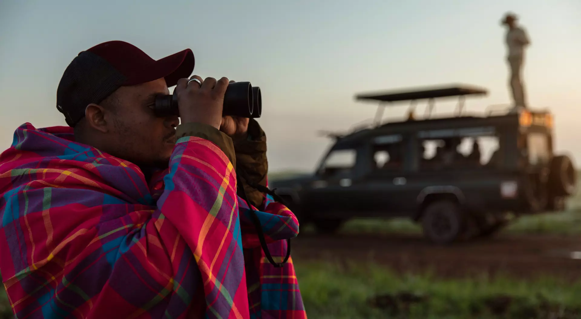 A Maasai safari tracker looks through binoculars while someone stands on a land cruiser in the background in Kenya.