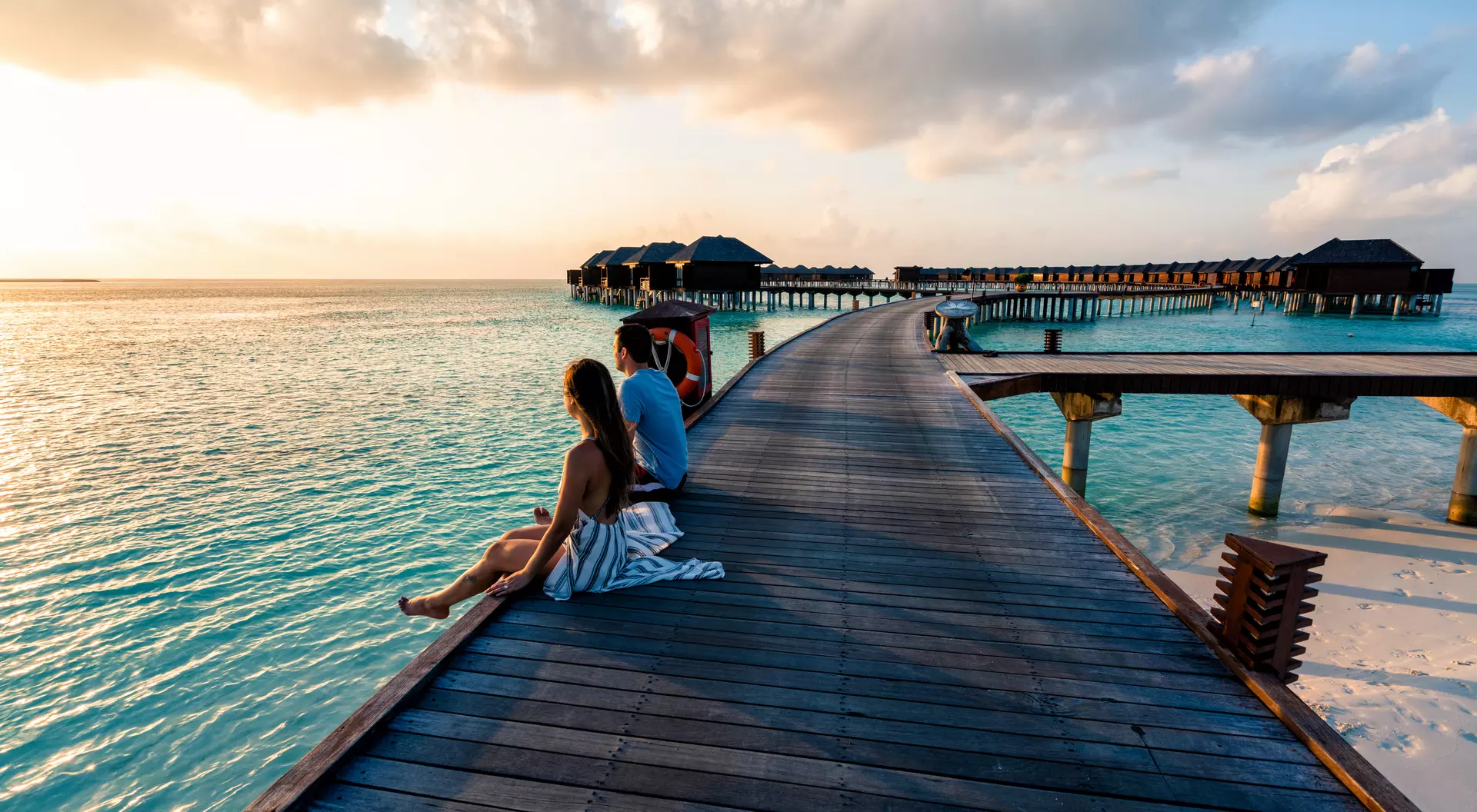 A couple sitting on a boardwalk leading out into the water in the Maldives.