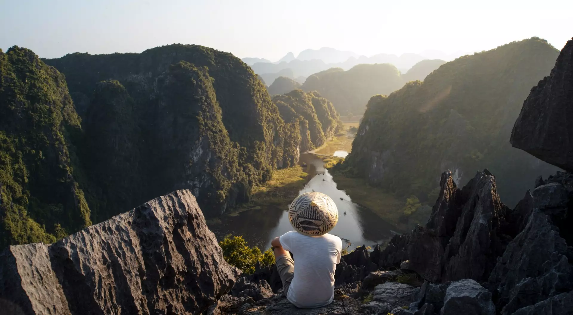 man in a straw hat sitting on the edge of a mountain overlooking river