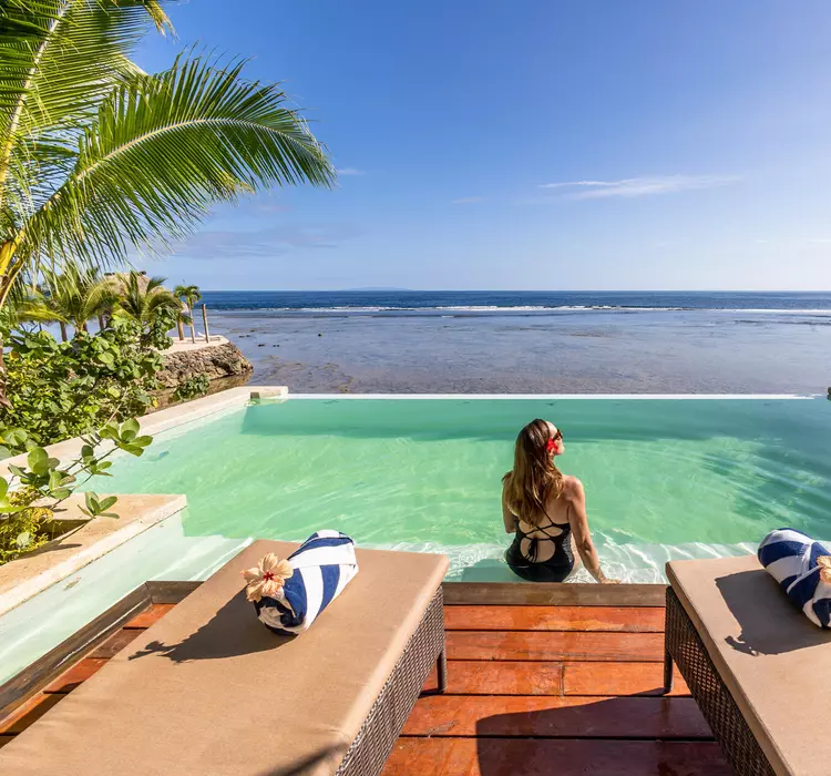 Woman lounging by a beach-side pool, surrounded by palm trees, clear, blue water and sunny skies.