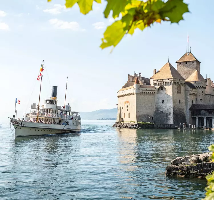 A vintage steamship sails near the historic Château de Chillon, a medieval castle on the shore of a clear lake, framed by green leaves.
