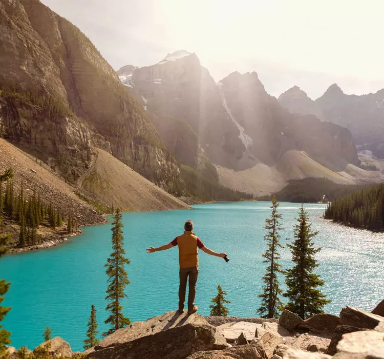 Person standing on the edge of rocks overlooking a clear blue lake with mountain backdrops.