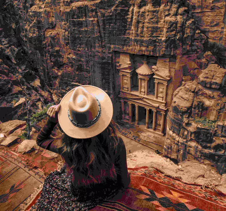 Woman with a hat sitting on the edge of a cliff overlooking a carved tomb