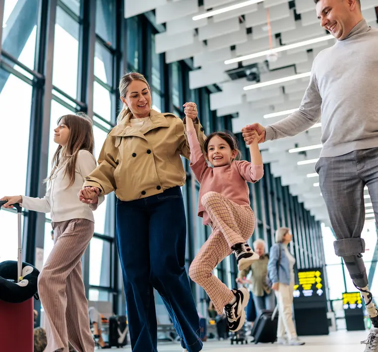 A cheerful family walks through a modern airport terminal with luggage before departure while the father uses a prosthetic leg. The parents hold hands with their daughters as they laugh and enjoy a playful travel moment surrounded by other travelers and airport activity.
