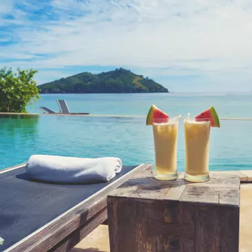ocktails by the pool with beach in the background. There are sun lounges with towels. The drinks are Pina coladas with fruit.