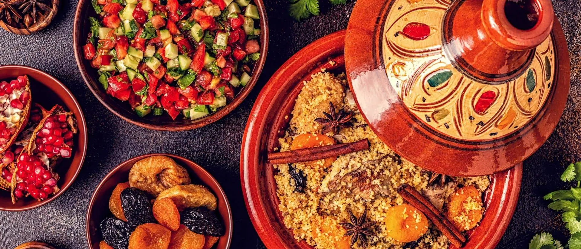 A colorful spread of couscous with spices, surrounded by salads, dried fruits, and garnishes on a textured surface.