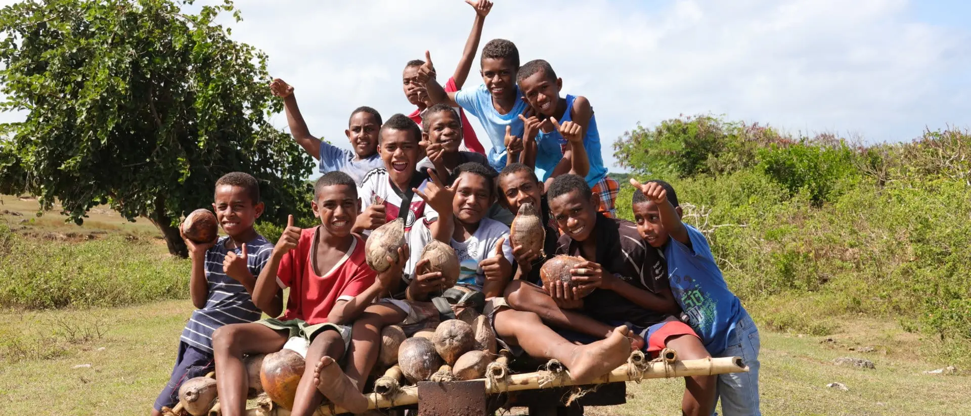 A group of joyful boys poses on a wooden cart, holding coconuts outdoors in a lush green setting.