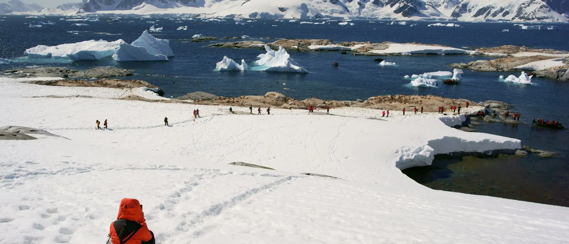 A person in a bright orange jacket sits on snowy terrain, overlooking a glacier and a group walking near the water.
