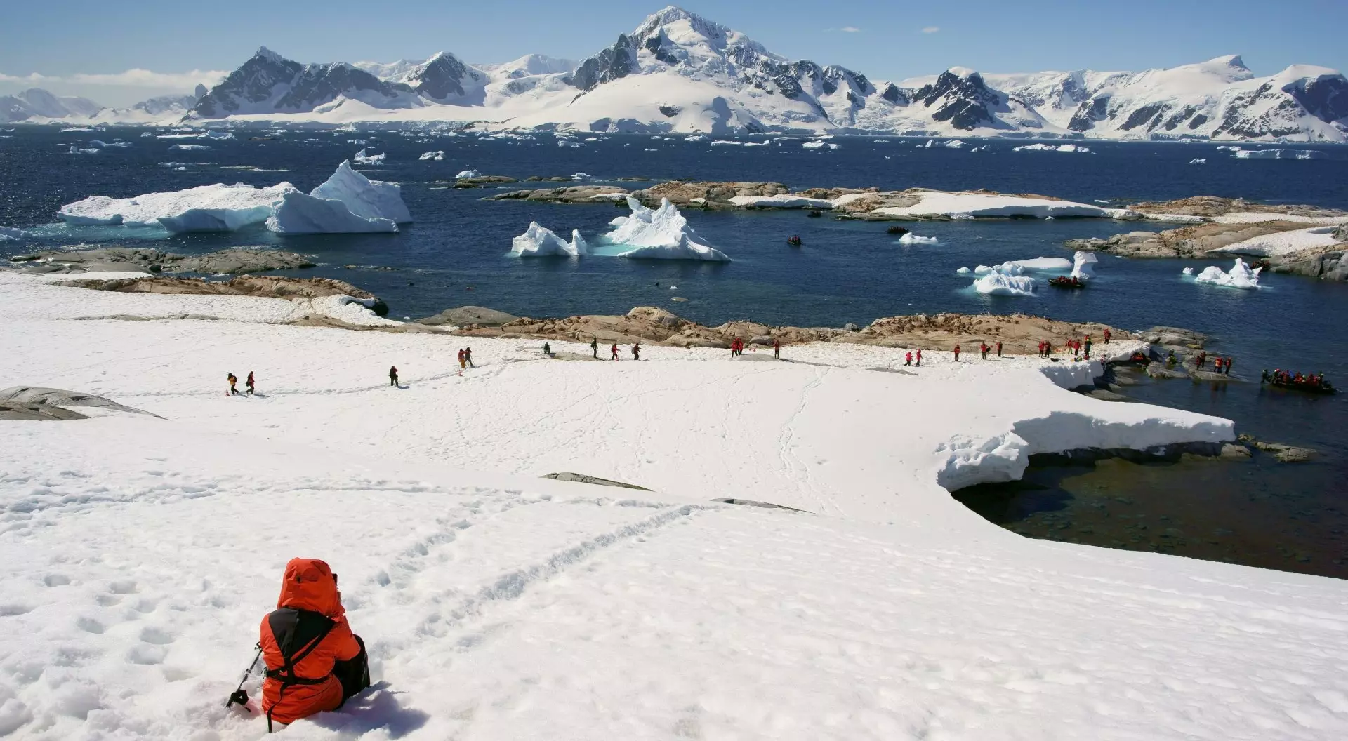 A person in a bright orange jacket sits on snowy terrain, overlooking a glacier and a group walking near the water.