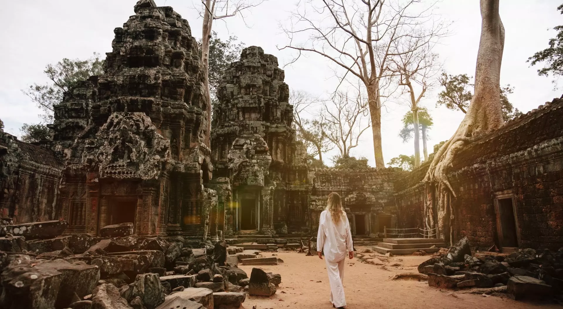 A person in white clothing walks through ancient temple ruins surrounded by trees and a sandy path.