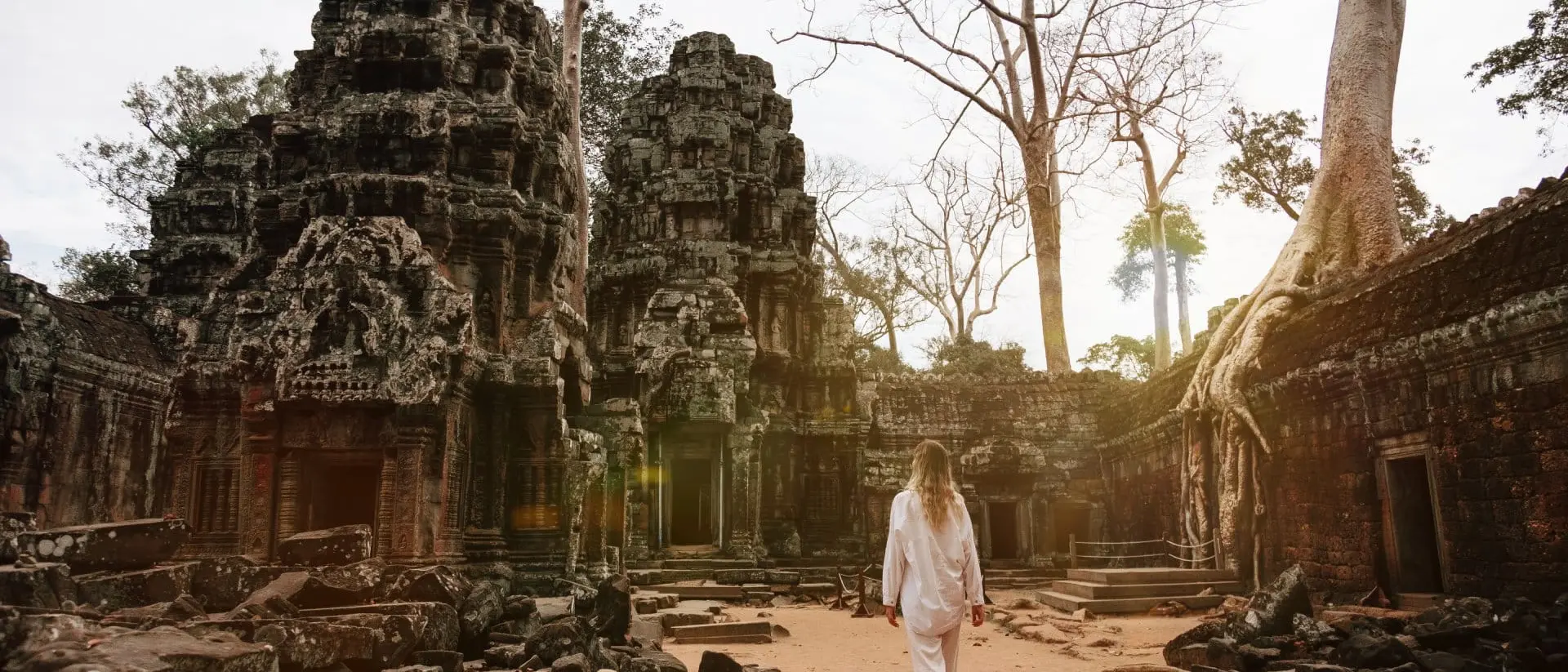 A person in white clothing walks through ancient temple ruins surrounded by trees and a sandy path.