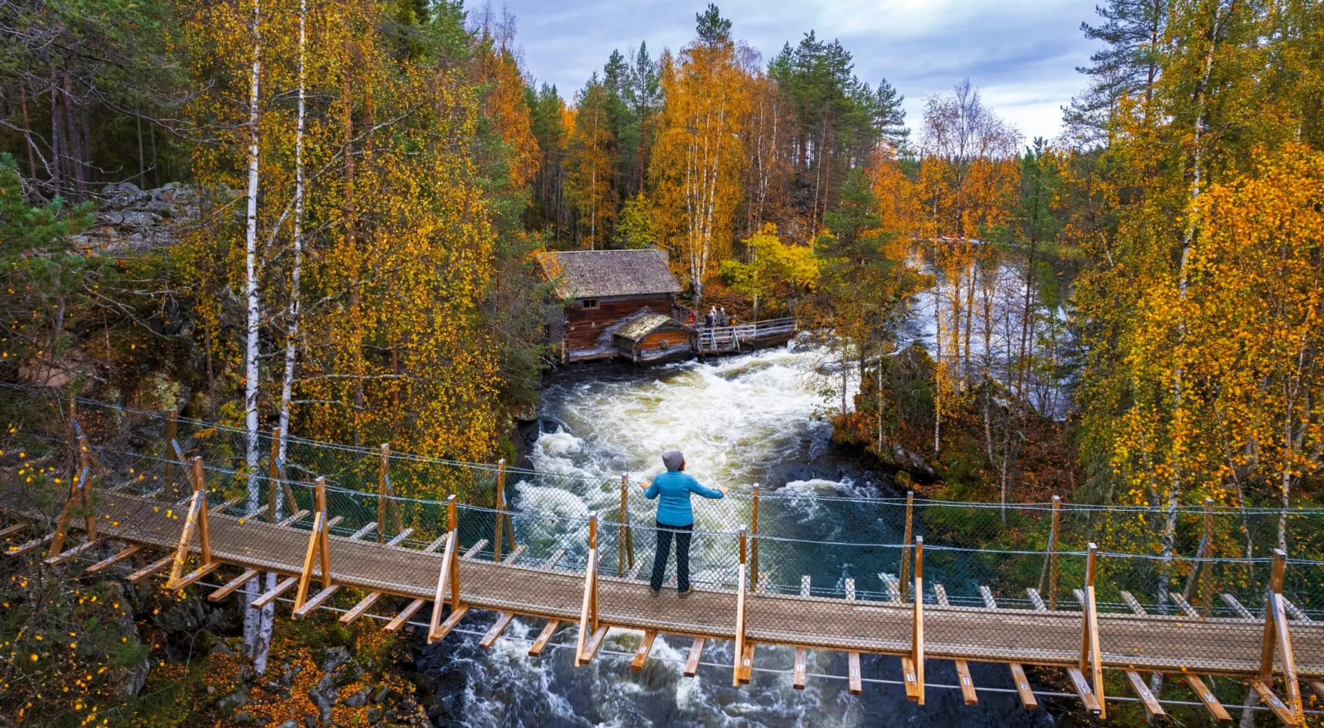 A person stands on a wooden bridge overlooking a river with autumn foliage and a cabin in the background.