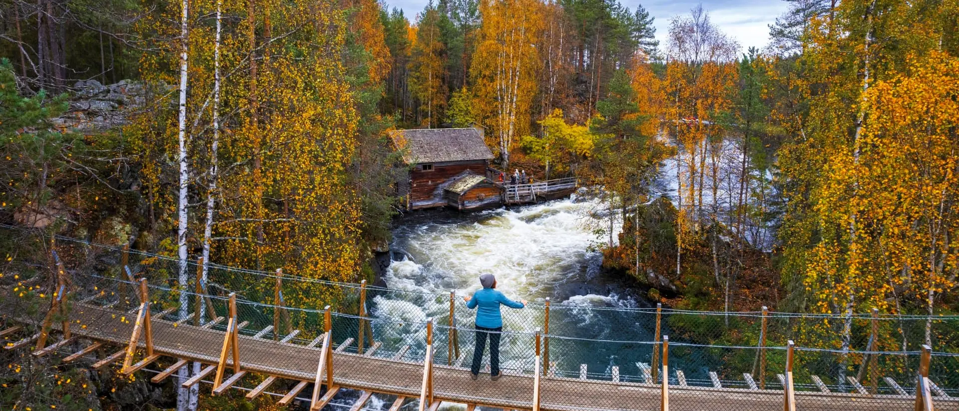 A person stands on a wooden bridge overlooking a river with autumn foliage and a cabin in the background.