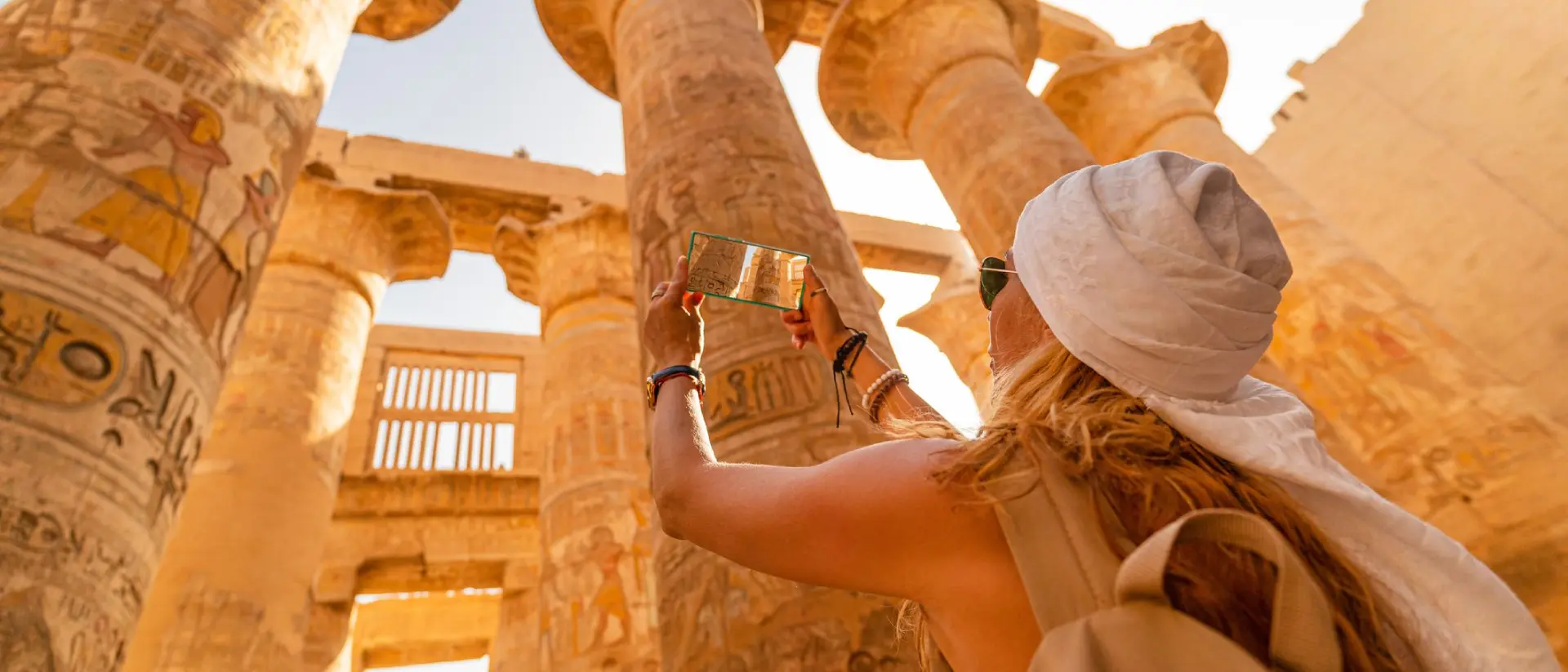 A person taking a photo of ancient columns with hieroglyphs in a sunlit archaeological site.