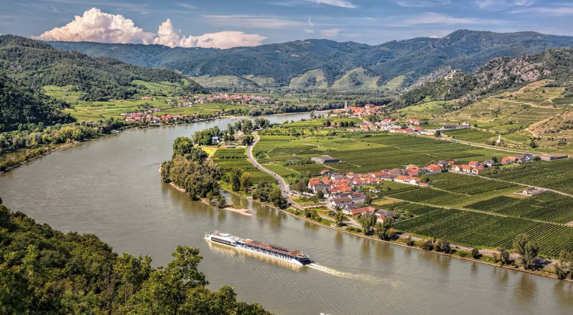 A scenic river winds through lush green hills and vineyards, with a boat navigating the water near a small village.