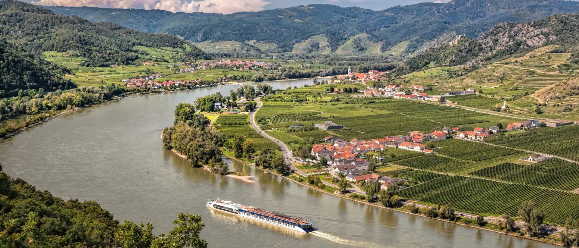 A scenic river winds through lush green hills and vineyards, with a boat navigating the water near a small village.
