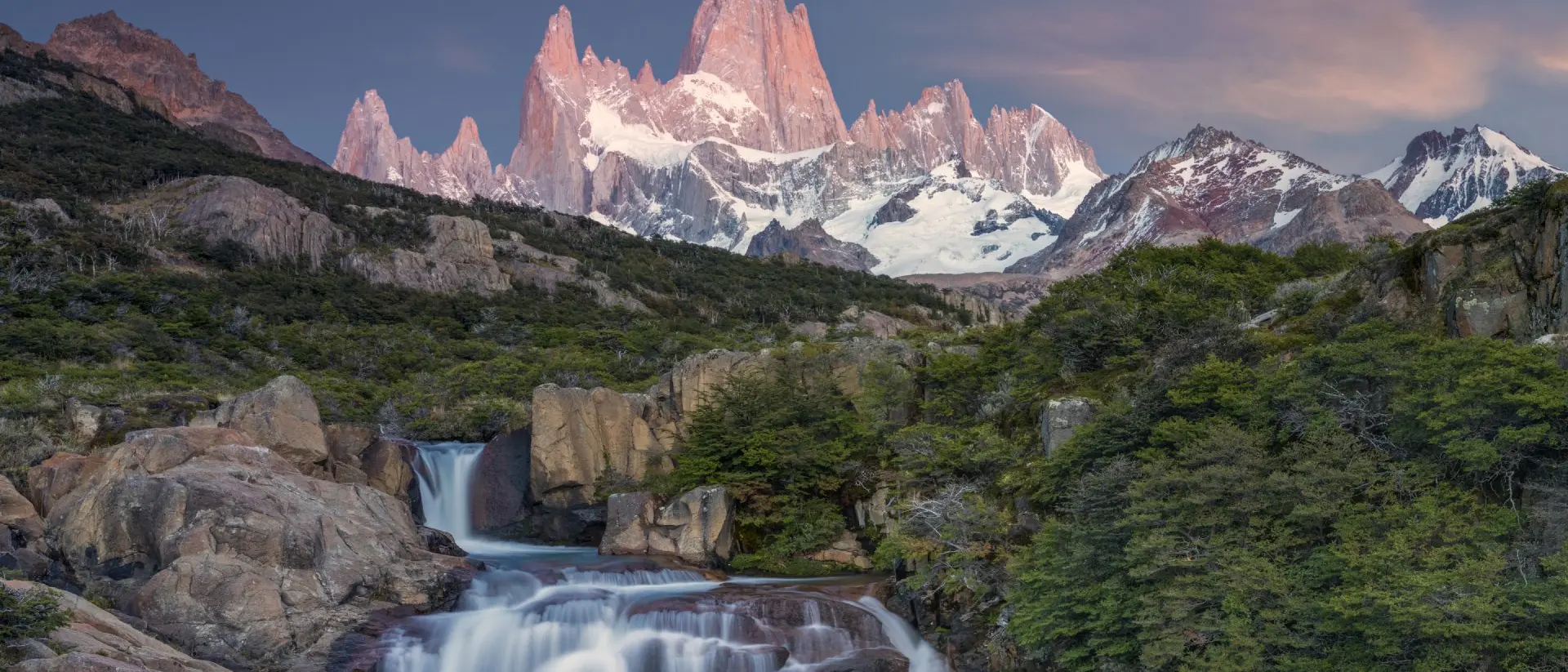 A serene landscape featuring a waterfall flowing through rocks, with majestic mountains and a colorful sky in the background.