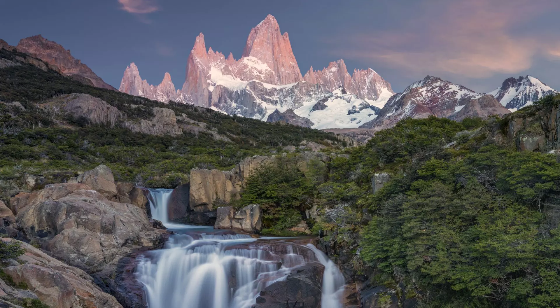 A serene landscape featuring a waterfall flowing through rocks, with majestic mountains and a colorful sky in the background.