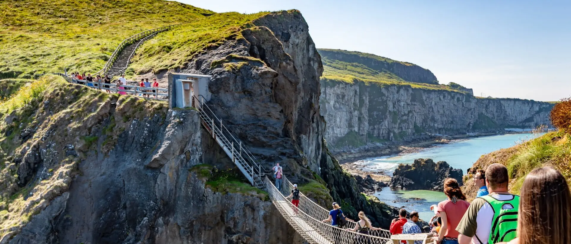 A sunny coastal scene showing a suspended bridge with people walking towards scenic cliffs and the ocean in the background.