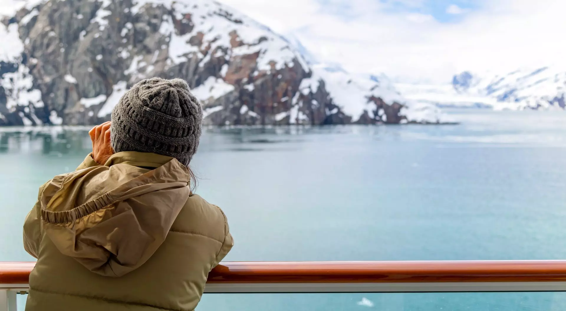 A young woman with binoculars views the snow covered mountains and glaciers from a cruise ship balcony at Glacier Bay National Park and Reserve, Alaska USA.