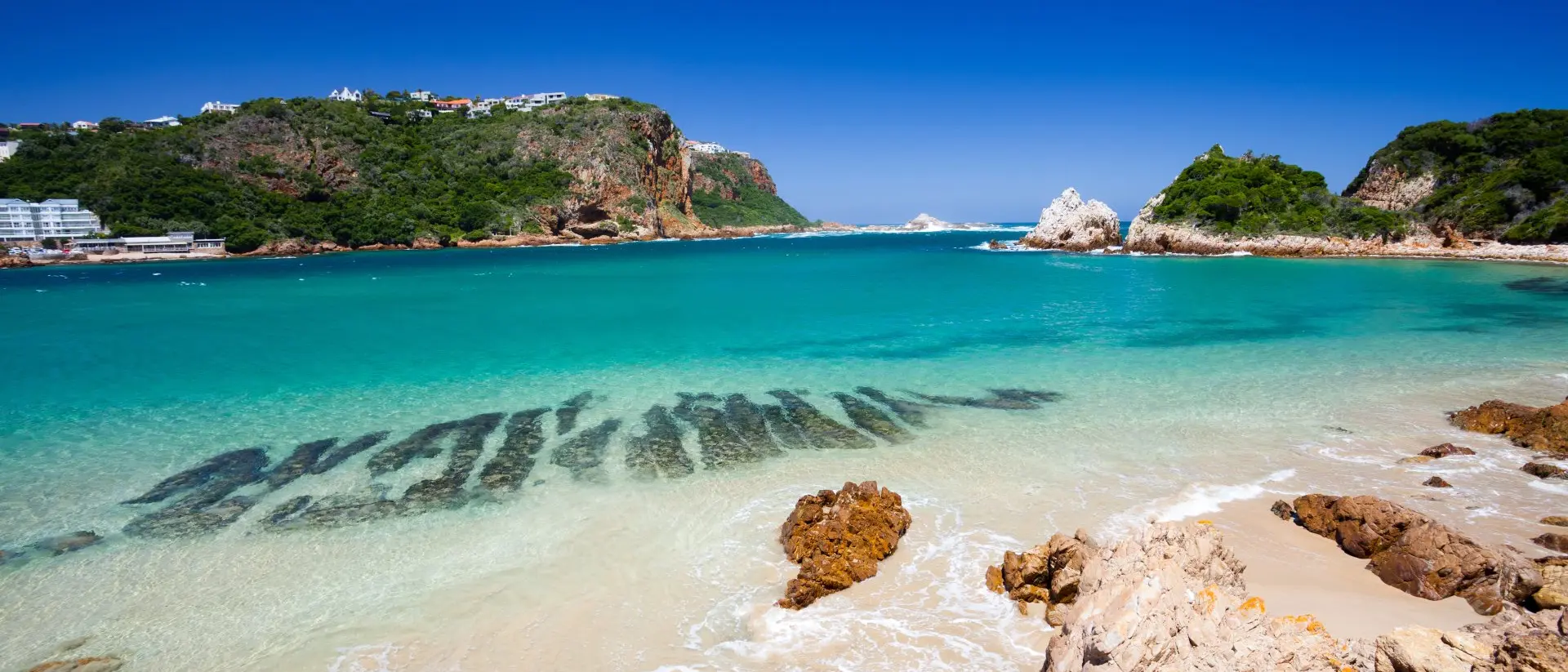 A beach in Knysna, Western Cape, South Africa. The beach is surrounded by lush green hills and has white sand and clear blue water.