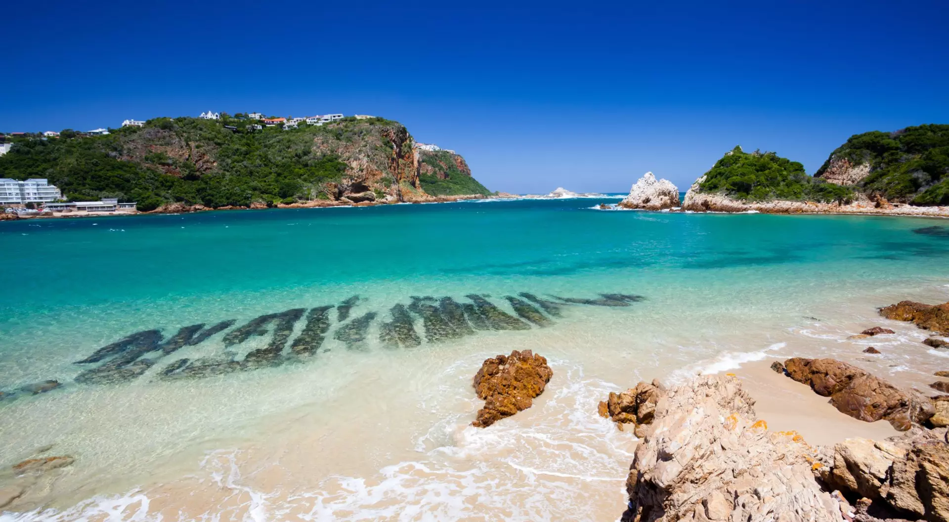 Knysna, Western Cape, South Africa A beach in Knysna, Western Cape, South Africa. The beach is surrounded by lush green hills and has white sand and clear blue water.