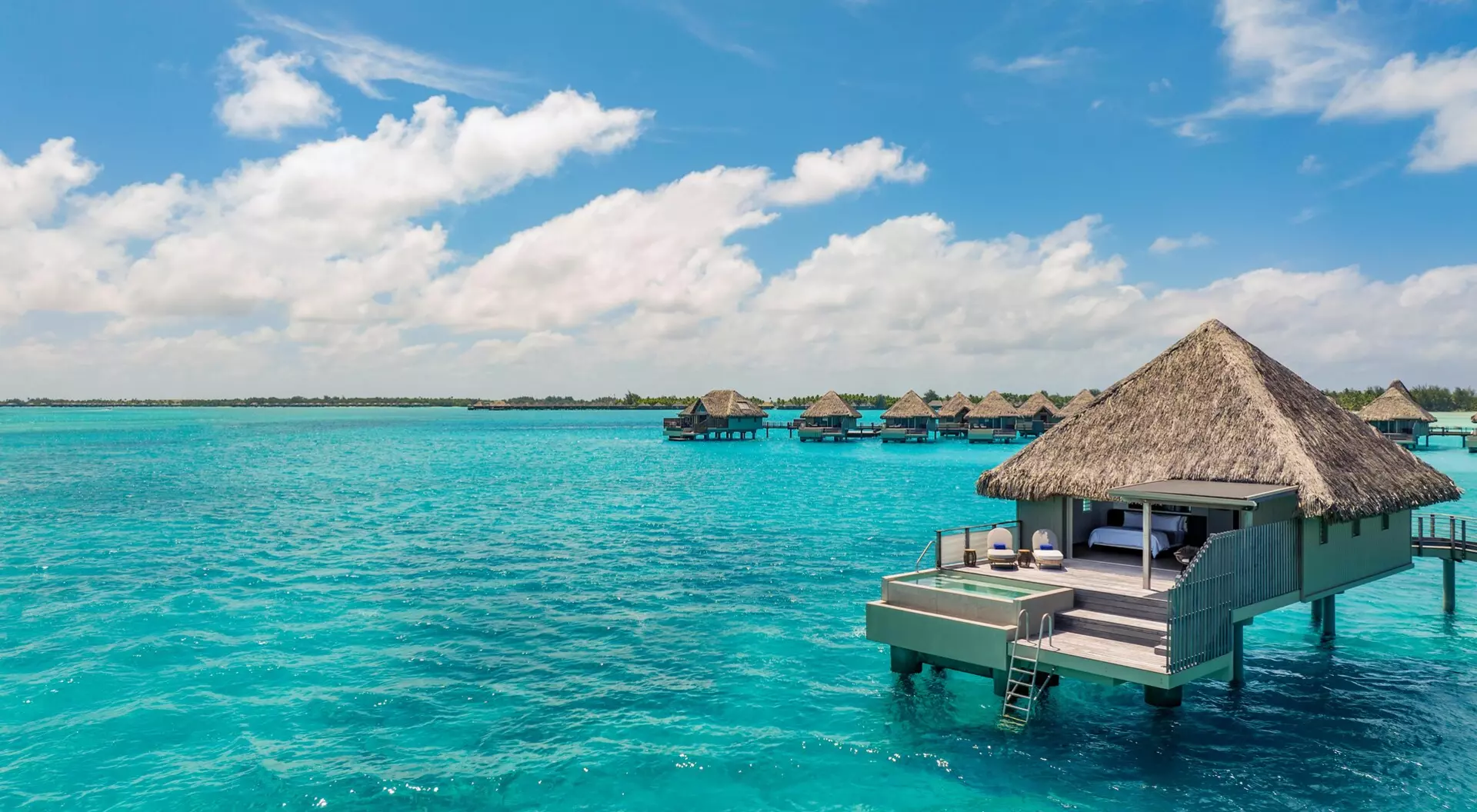 Overwater bungalows with thatched roofs sit above bright turquoise water under a sunny sky, evoking a luxurious tropical island getaway.
