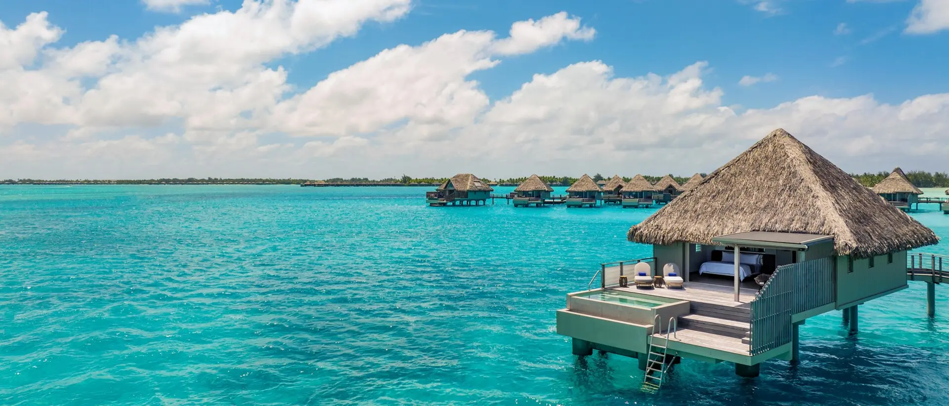 Overwater bungalows with thatched roofs sit above bright turquoise water under a sunny sky, evoking a luxurious tropical island getaway.
