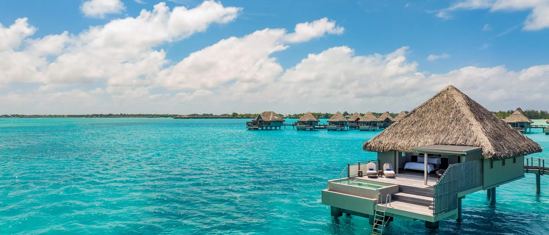 Bora Bora, French Polynesia Overwater bungalows with thatched roofs sit above bright turquoise water under a sunny sky, evoking a luxurious tropical island getaway.
