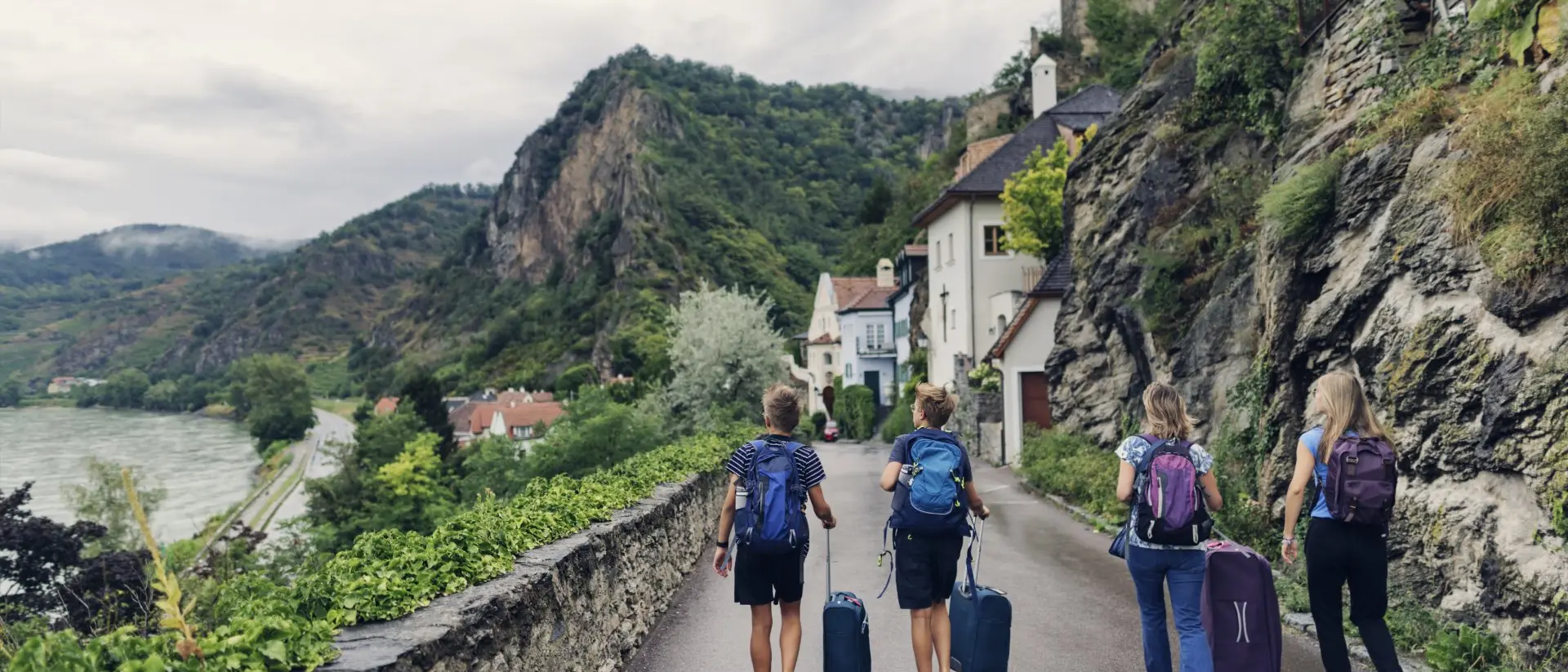 Travelers with suitcases walk along a scenic road surrounded by mountains and greenery near a riverbank.