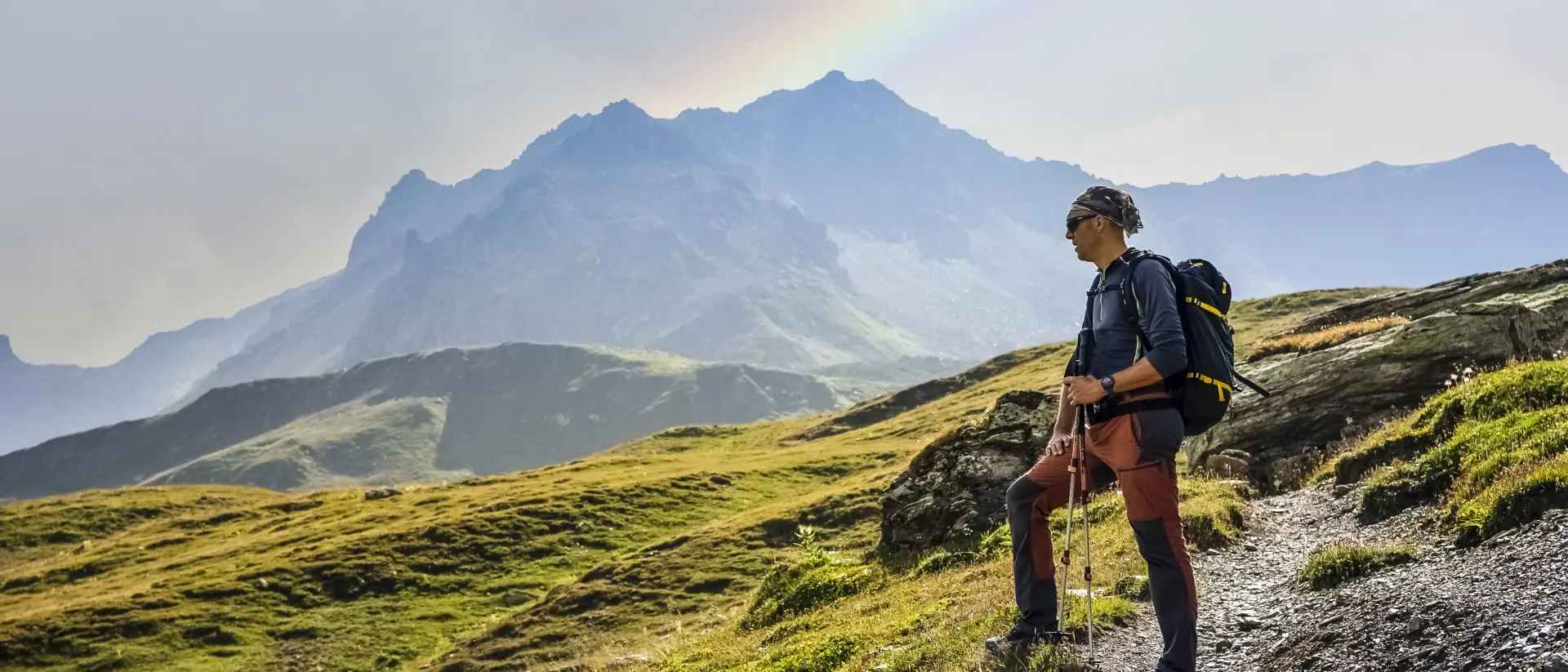 Argentina Hiker standing on a rocky trail, gazing at mountains and a rainbow in the sky.