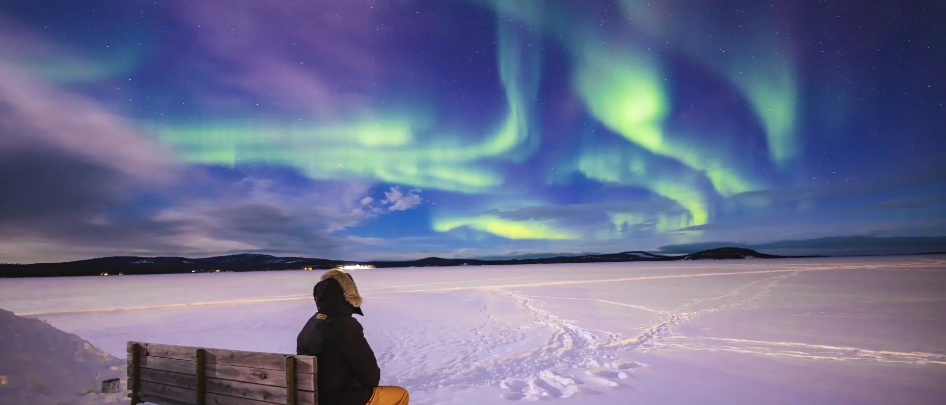 Person sitting on a bench, gazing at vibrant northern lights over a snowy landscape.