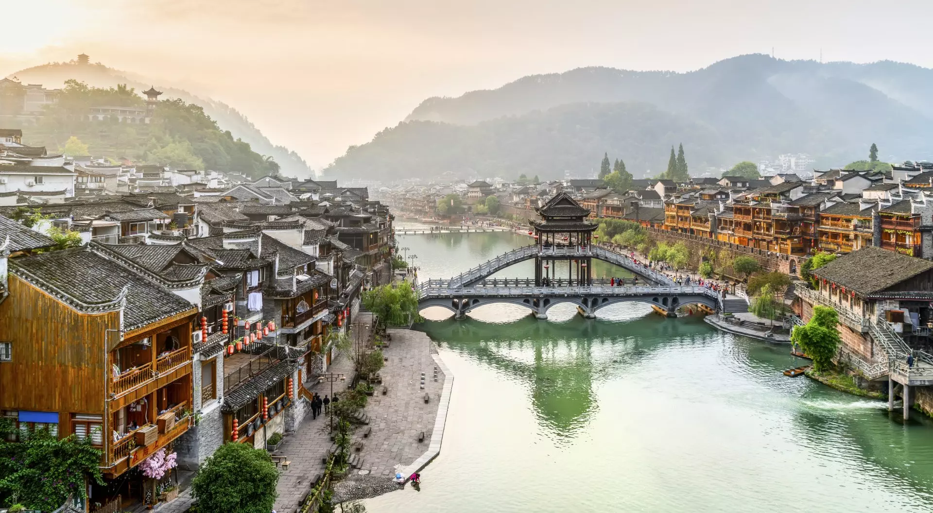 Scenic view of a river lined with traditional wooden buildings, a bridge, and mountains in the background at sunset.
