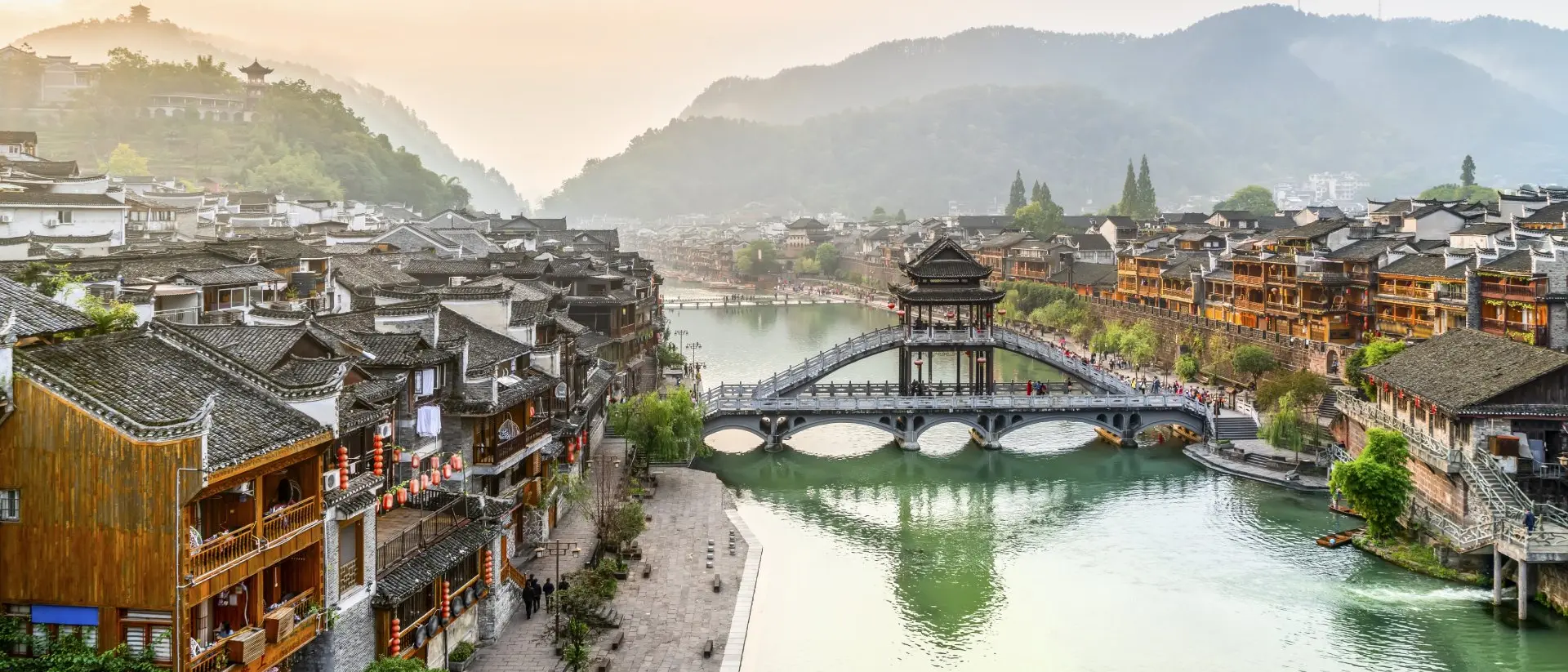 Scenic view of a river lined with traditional wooden buildings, a bridge, and mountains in the background at sunset.