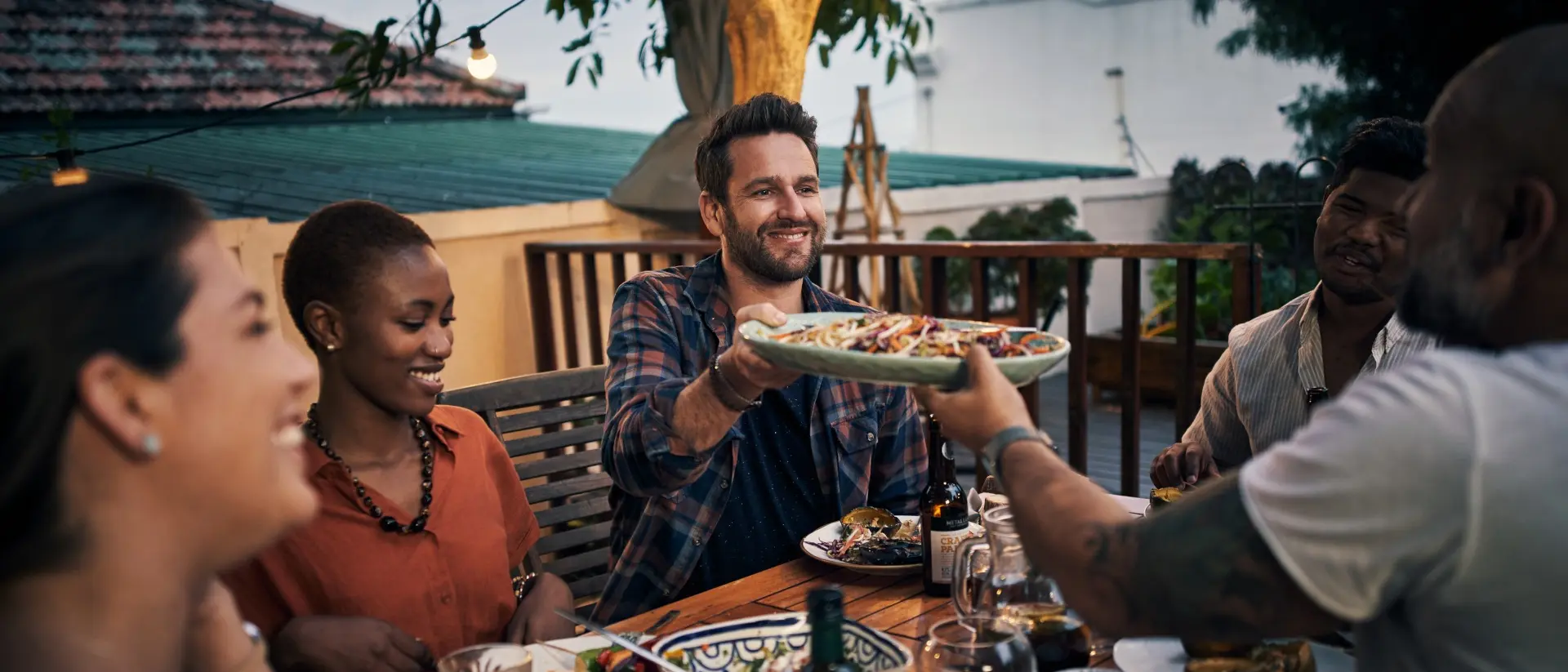 A group of friends enjoys dinner outdoors, sharing food and smiling at each other under string lights.