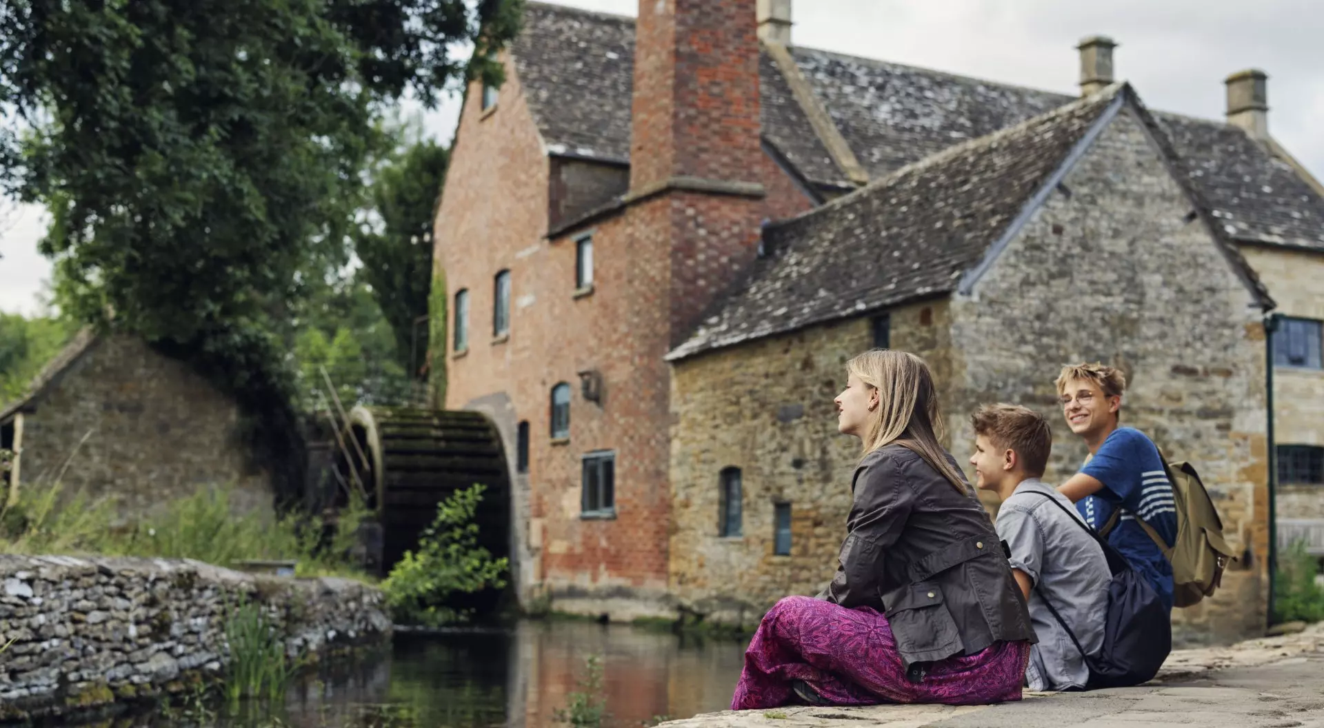 Three children sit by a river, enjoying the view of a rustic building and a waterwheel in a peaceful countryside setting.