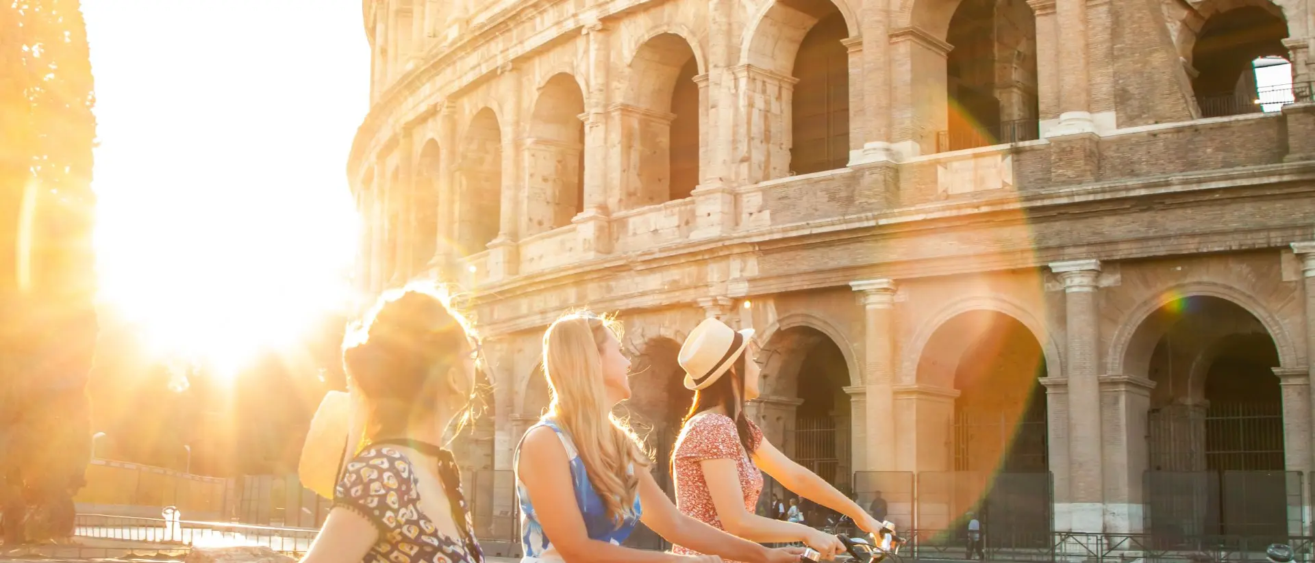 Three happy young women friends tourists with bikes at Colosseum in Rome, Italy at sunrise