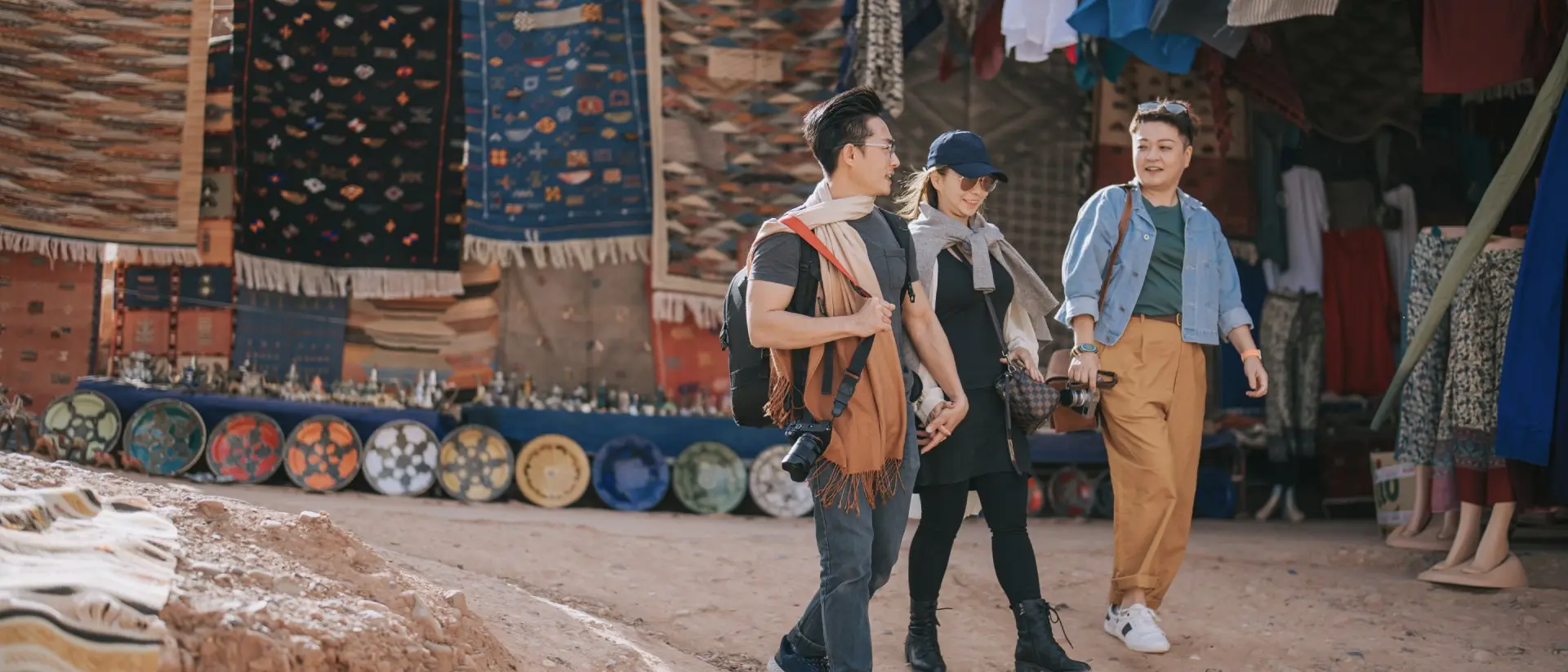 Three people walk through a vibrant market, surrounded by colorful textiles and handmade pottery.