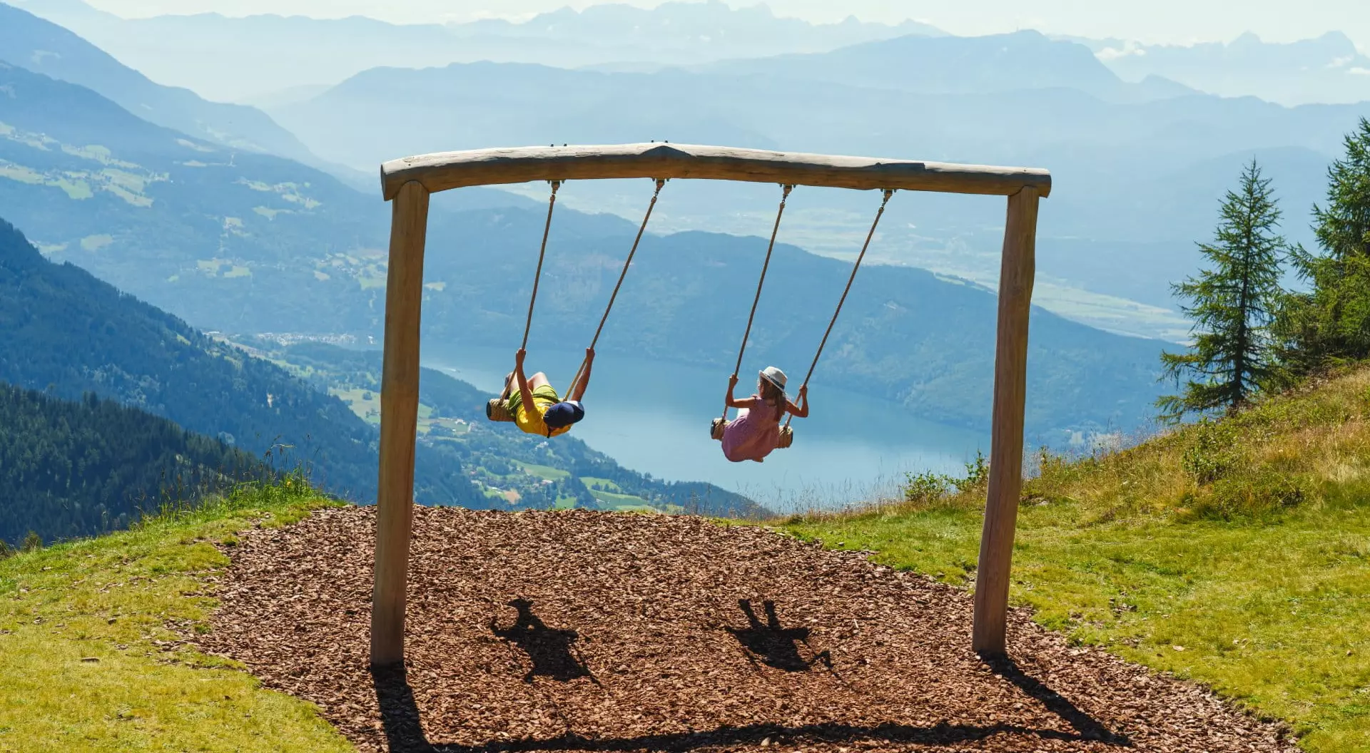 Two children swing on a wooden swing set high in the mountains, with a scenic view of valleys and distant peaks.