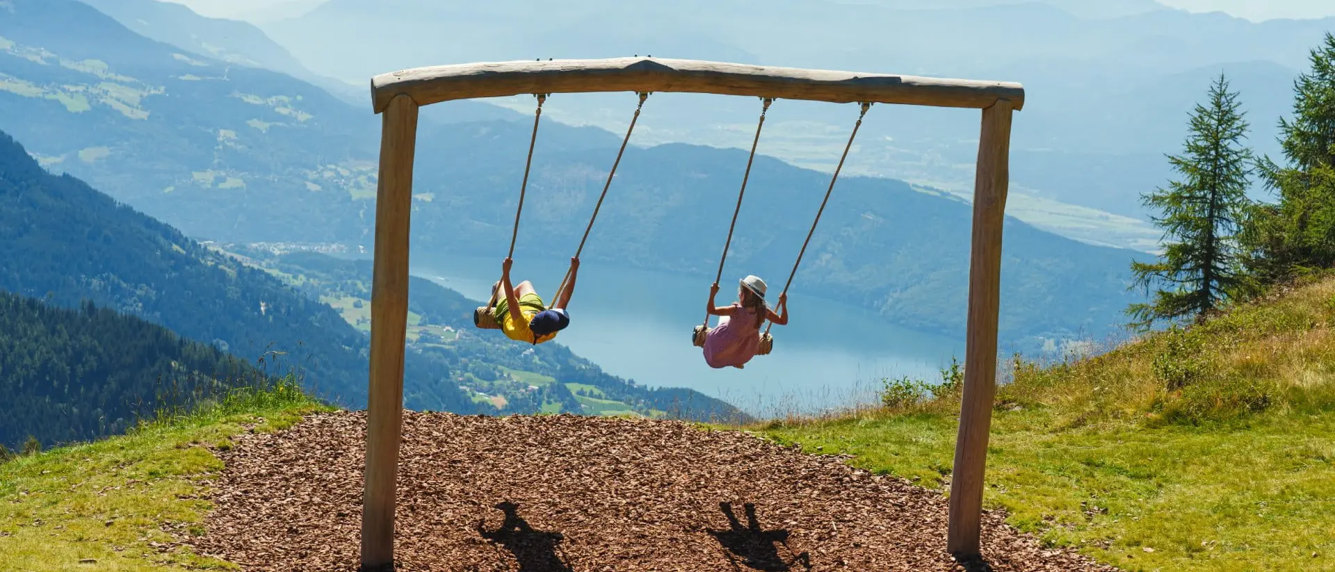 Two children swing on a wooden swing set high in the mountains, with a scenic view of valleys and distant peaks.