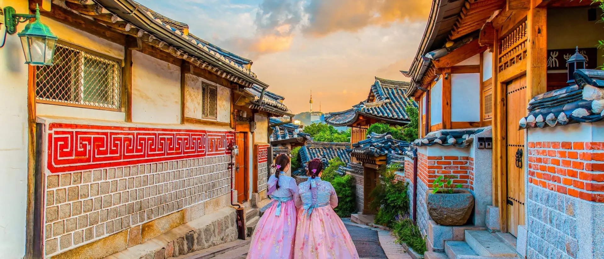 Two girls in traditional attire stroll through a picturesque alley lined with wooden houses and colorful sky at sunset.